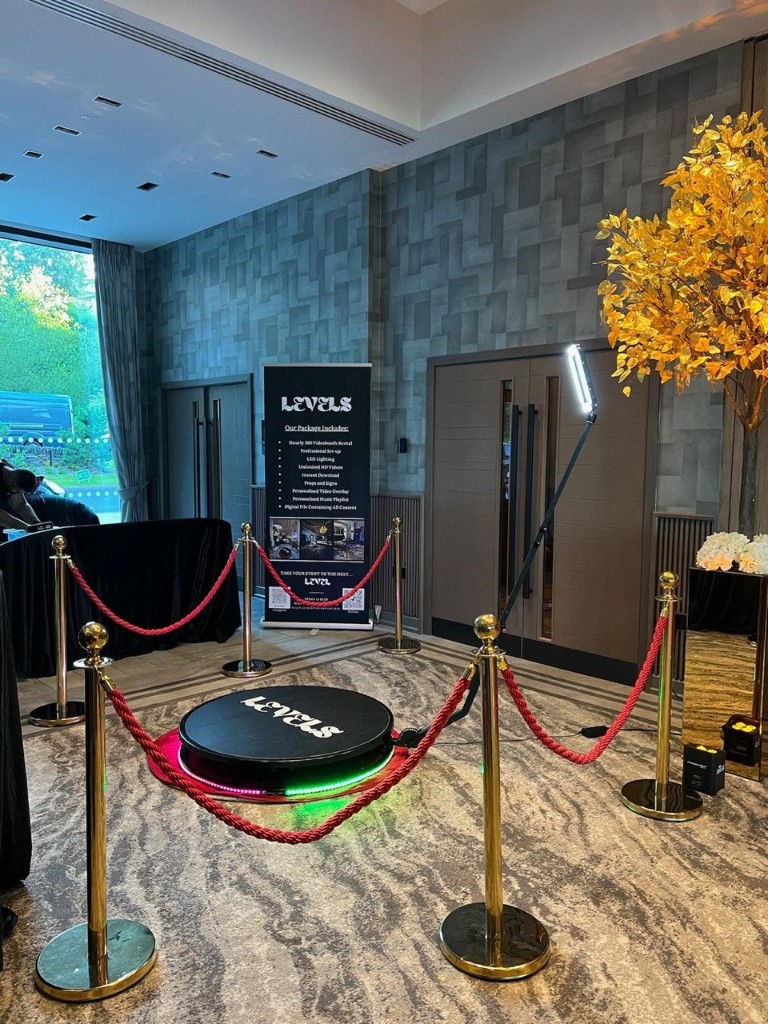 360 Video Booth with LEVELS platform, LED base, gold stanchions, red ropes, package banner, and gold-leaf tree by floor-to-ceiling windows