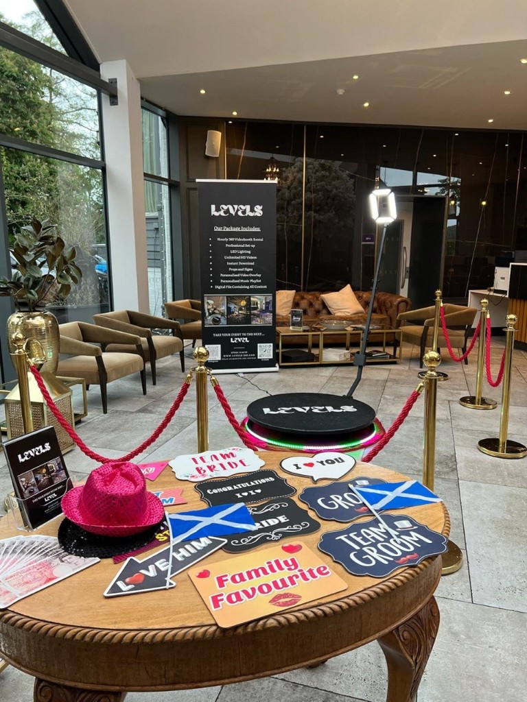 Props table with signs and hats beside the 360 platform, package banner, and VIP ropes in a bright lounge with sofas and windows