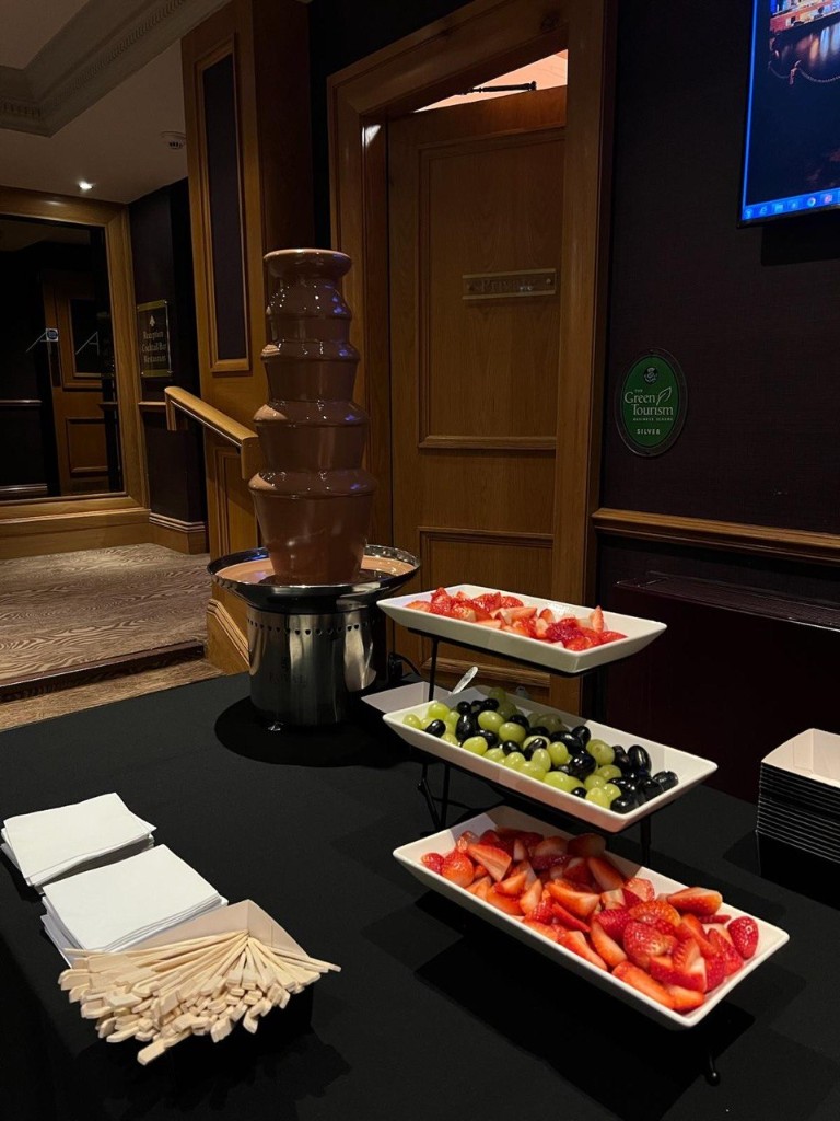 Five-tier milk chocolate fountain with three-tier stand of strawberries and grapes on a black-clothed table